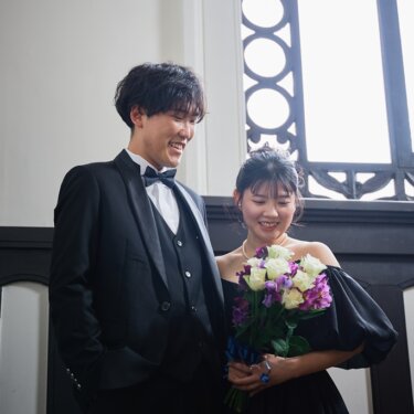 横浜開港記念会館にて、装飾的な鉄格子窓を背に、白と紫のブーケを見つめて笑う新郎新婦。クラシカルなウェディングフォト