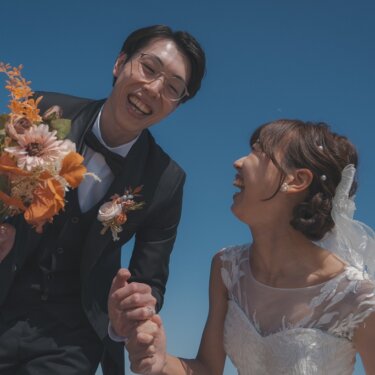 青空の下、ブーケを掲げて笑う新郎と、見上げて大笑いする花嫁。ローアングルで撮る楽しいウェディングフォト