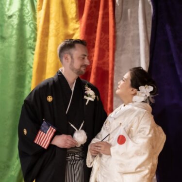久良岐公園にて、五色の揚幕を背景に、日本とアメリカの国旗を持って見つめ合う新郎新婦。国際結婚の和装前撮り
