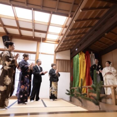 久良岐能舞台の揚幕の前に立つ新郎新婦を、和室から拍手で祝福する家族。温かな和装フォトウェディング
