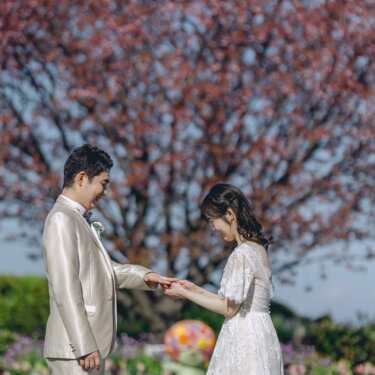 赤い花の木とカラフルな花壇を背景に、指輪を確かめ合う新郎新婦。レースのウェディングドレスで撮る前撮り