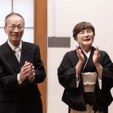 障子の前で拍手して祝福する、スーツの父と黒留袖の母。和装前撮りに立ち会う家族のスナップ