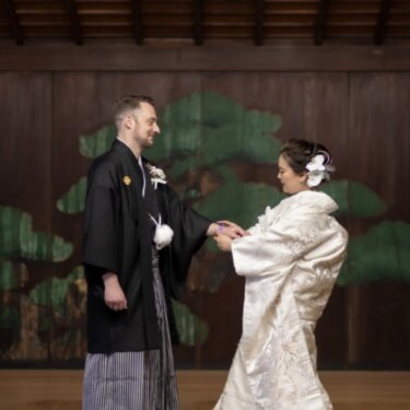 横浜の久良岐能舞台で、松の鏡板の前で手を取り合い見つめ合う紋付袴の新郎と白無垢の花嫁の和装前撮り