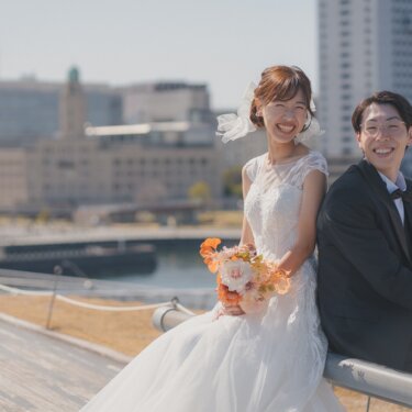 大さん橋の柵に並んで腰を下ろし、港の街並みを背に笑う新郎新婦。オレンジのブーケが映えるウェディングフォト