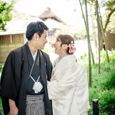 竹林の中で頬を寄せて笑い合う黒紋付袴の新郎と白無垢の花嫁。茅葺屋根が見える横溝屋敷の和装前撮り