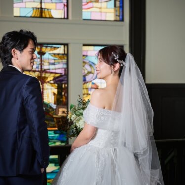 帆船のステンドグラスの前で振り返って笑う、ベールの花嫁と新郎の後ろ姿を写した洋館の結婚写真