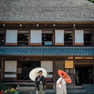 茅葺と瓦の二層屋根が特徴の横溝屋敷の母屋を背に、白と朱赤の和傘を持って並ぶ新郎新婦。和装ロケーションフォト