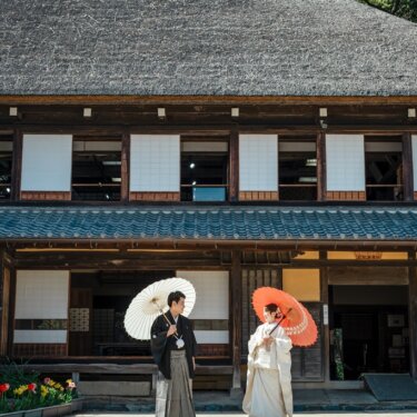 横溝屋敷の茅葺屋根の母屋とチューリップの花壇を背に、和傘を差して立つ新郎新婦の引きの構図の結婚写真