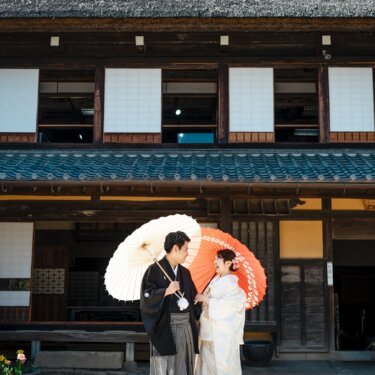 横溝住宅にて、茅葺屋根の古民家の前で、白と朱赤の和傘を差して向かい合う紋付袴の新郎と白無垢の花嫁の和装前撮り