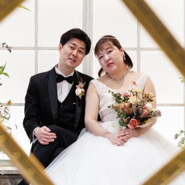 金の格子の枠越しに、窓辺に並んで座る新郎新婦を写したフレーム構図のスタジオフォト