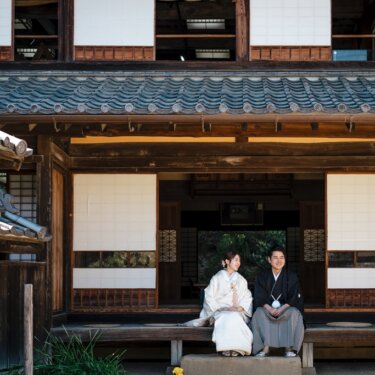 横溝屋敷の母屋の縁側に並んで座る新郎新婦を、瓦屋根と障子の建物ごと写した和装フォトウェディング