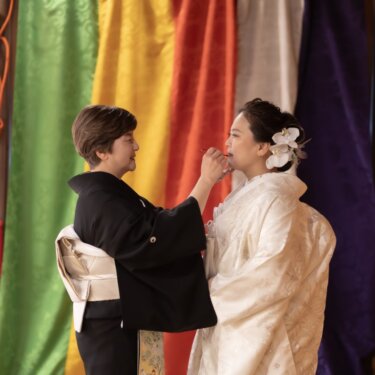 久良岐能舞台の五色の揚幕の前で、黒留袖の母が白無垢の花嫁の口紅を直す一場面。家族で撮る和装前撮り