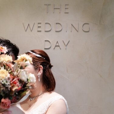 『THE WEDDING DAY』の立体文字が飾られた壁の前で、大きなブーケで顔を寄せ合う新郎新婦のスタジオフォト