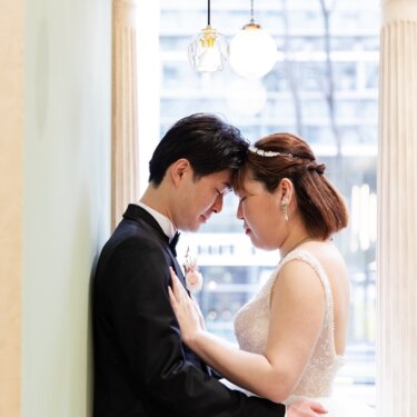 アーチ窓の柱とペンダントライトを背景に、額を寄せ合って抱き合う新郎新婦。ウェディングドレスで撮るロマンチックな前撮り