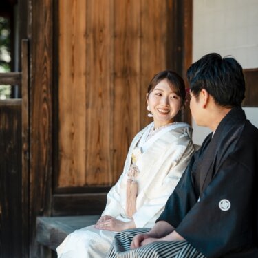 木の引き戸の前の縁側に並んで座り、笑い合う白無垢の花嫁と紋付袴の新郎の和装フォトウェディング