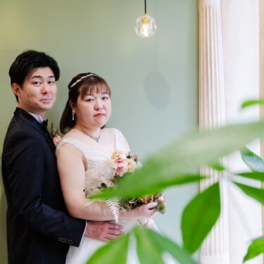 観葉植物を前景にぼかし、ミントグリーンの壁の前でカメラを見つめる新郎新婦。ナチュラルなスタジオフォト