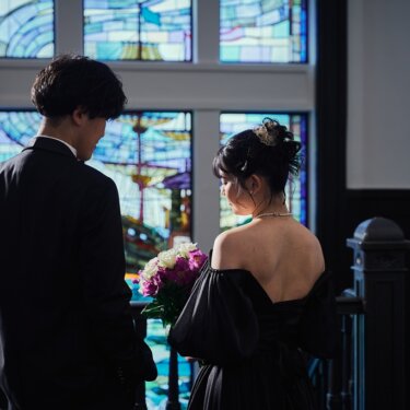 横浜開港記念会館にて、ステンドグラスの前で向かい合う新郎新婦の後ろ姿。背中の開いた黒いカラードレスが映える前撮り