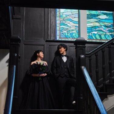 横浜開港記念会館にて、海と帆船のステンドグラスを背に、階段の踊り場に並んで立つ黒いドレスの花嫁とタキシードの新郎の結婚写真