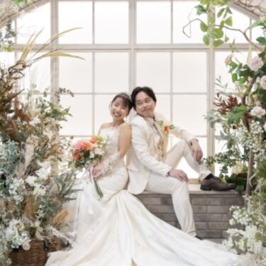 大きなアーチ窓と花々を望む窓辺に座る新郎新婦を正面から写した、リゾート風のスタジオフォト