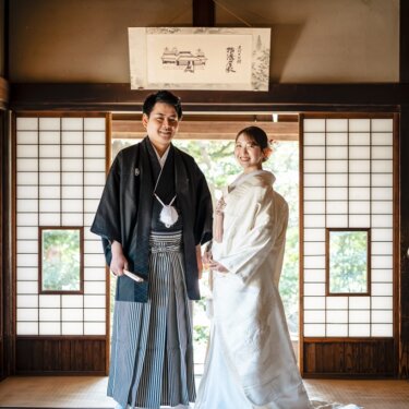 掛け軸と障子のある座敷に並んで立つ新郎新婦の全身ショット。古民家で撮る和装の結婚写真
