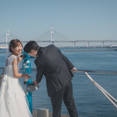 横浜ベイブリッジと海を背に、双眼鏡型の望遠鏡をのぞいて笑い合う新郎新婦。遊び心のあるロケーションフォト
