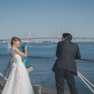 ベイブリッジを望む柵際で、望遠鏡をのぞいて笑い合う新郎新婦の後ろ姿。大さん橋で撮る結婚写真
