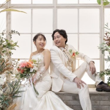 ドライフラワーと花に囲まれたアーチ窓のセットで、窓辺に腰かけて笑い合う新郎新婦のスタジオ前撮り