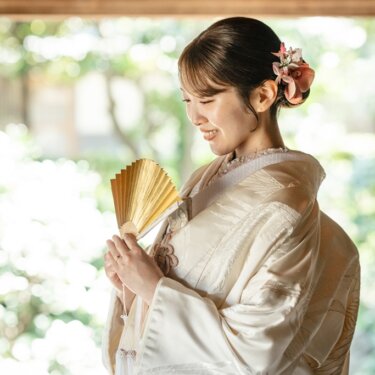 横溝住宅にて、金の扇子を手にほほえむ白無垢の花嫁の横顔。庭の緑をぼかした和装前撮りのソロショット