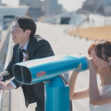 望遠鏡をのぞく花嫁と、手すりにもたれて笑う新郎。みなとみらいの街並みを望むウェディングフォト