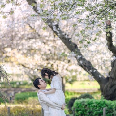 大きな桜の木の下で新郎が花嫁を高く抱き上げる、引きの構図の春のフォトウェディング