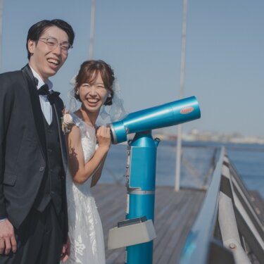 大さん橋の青い望遠鏡の前で、大笑いするタキシードの新郎とベールの花嫁。遊び心ある横浜の前撮り