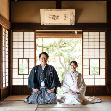 掛け軸の下の座敷に並んで正座し、ほほえむ紋付袴の新郎と白無垢の花嫁。古民家で撮る結婚写真