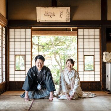 床の間のある古民家の座敷に正座し、カメラに笑顔を向ける黒紋付袴の新郎と白無垢の花嫁の和装前撮り
