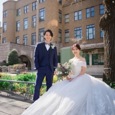 歴史ある近代建築を背に、花壇のそばでカメラに笑顔を向けるネイビーのタキシードの新郎とウェディングドレスの花嫁の前撮り
