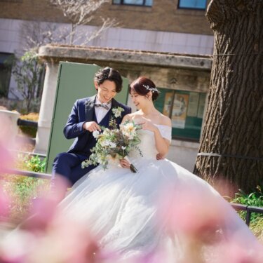 ピンクの花を前景にぼかし、花壇に座ってブーケを見つめ笑い合う新郎新婦のウェディングフォト