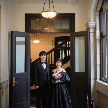 横浜市開港記念会館の木の扉の前に並んで立つ、タキシードの新郎と黒いドレスの花嫁の全身を写した前撮り
