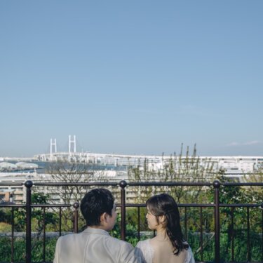 港の見える丘公園のベンチに並んで座り、ベイブリッジを眺める新郎新婦の後ろ姿。肩を抱く新郎がやさしい前撮り