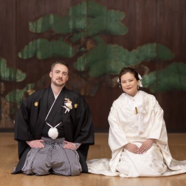 久良岐能舞台の松が描かれた鏡板を背に正座し、カメラを見つめる新郎新婦。格式ある和装の結婚写真