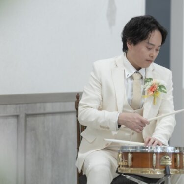 真剣な表情でスネアドラムを叩く白いタキシードの新郎のソロショット。音楽好きの二人らしいフォトウェディング