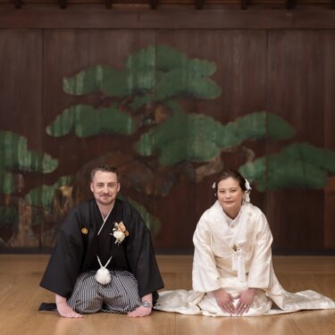 久良岐能舞台の松の鏡板の前に正座する紋付袴の新郎と白無垢の花嫁を、引きで写した和装前撮り