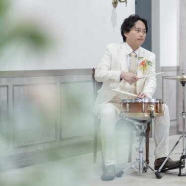 観葉植物を前景にぼかし、スネアドラムを叩く白いタキシードの新郎を写したスタジオフォト。趣味を取り入れた前撮り
