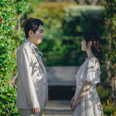 緑のアーチに囲まれたローズガーデンの小道で、少し離れて見つめ合う新郎新婦。ナチュラルなレースドレスのフォトウェディング