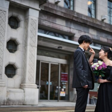 石造りの建物の前で、花嫁が新郎の鼻をつまむほほえましい一枚。黒いドレスで撮る遊び心ある前撮り