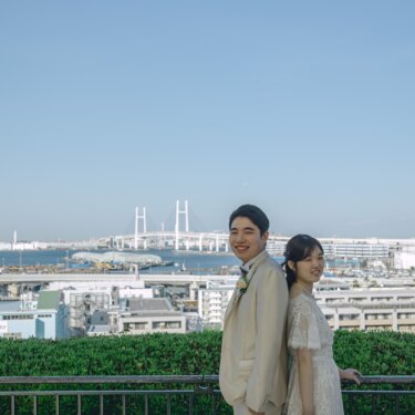 港の見える丘公園の展望台で、ベイブリッジを背に背中合わせで立つベージュのタキシードの新郎とレースのウェディングドレスの花嫁の前撮り