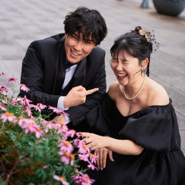 花壇のそばに腰を下ろし、指をさし合って大笑いする新郎新婦。黒いオフショルダーのドレスで撮る結婚写真