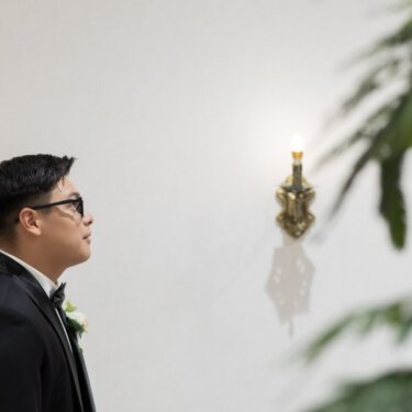 観葉植物をぼかし、壁灯の前で視線を上げる新郎の横顔。ファーストミート前のスタジオフォト
