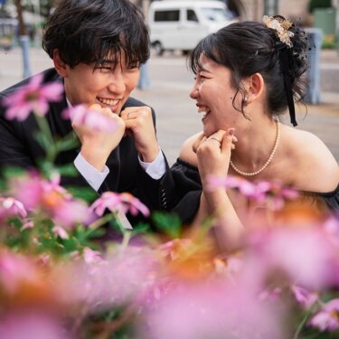 ピンクの花壇越しに向かい合い、両手を握って笑い合う新郎新婦。黒いカラードレスで撮る街ロケの前撮り