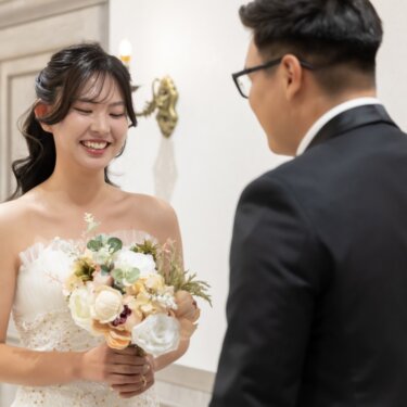 新郎の後ろ姿越しに、ブーケを持って照れ笑いする花嫁。ファーストミートで撮る前撮り