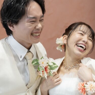 テラコッタ色の壁を背に、はじけるように大笑いする新郎新婦のアップ。ブートニアの花が映える自然体のウェディングフォト