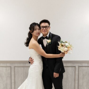 壁灯のある白い壁の前で抱き合い、頬を寄せてカメラにほほえむ新郎新婦のウェディングフォト